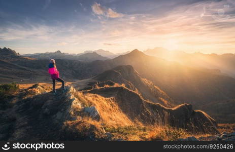 Young woman on mountain peak and beautiful mountain valley in haze at colorful sunset in autumn. Dolomites, Italy. Sporty girl, mountain ridges in fog, orange grass, trees, golden sun in fall. Hiking	