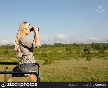 Young woman on safari standing in jeep looking through binoculars side view