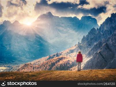 Young woman on the hill is looking on the majestic mountains at sunset in autumn in Dolomites, Italy. Landscape with girl, cloudy sky, sunbeam, high rocks and trees in fall. Travel in italian alps