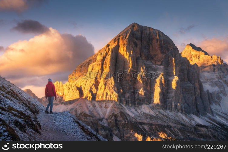 Young woman on the trail looking on high mountain peak at sunset in Dolomites, Italy. Autumn landscape with girl, path, rocks, sky with clouds at colorful sunset. Hiking in alps. Majestic mountains