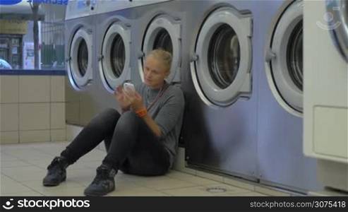 Young woman passing the time with cellphone while waiting for the laundry. She sitting on the floor by washing machines