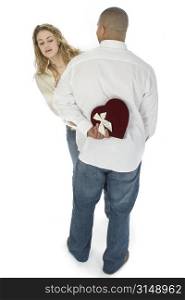 Young woman peeking around man&acute;s back to see what he&acute;s holding. Heart shaped box of chocolates.