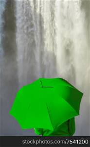 Young woman person in a green coat or jacket holding a green umbrella in front of a waterfall