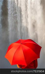 Young woman person in a red coat or jacket holding a red umbrella in front of a waterfall