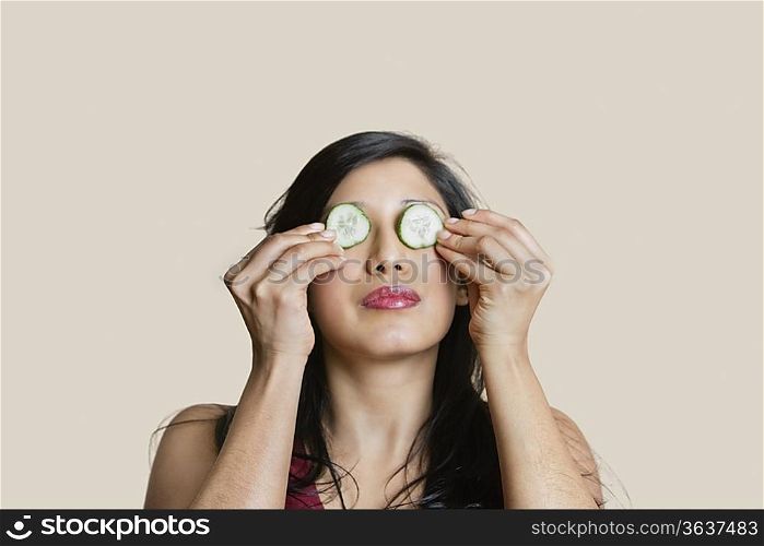 Young woman placing cucumber on eyes over colored background