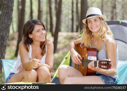 young woman playing guitar in the campsite while friend listening