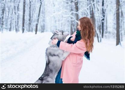 Young woman playing with siberian husky, snowy forest on background. Cute girl with playful charming dog. Female person funs with domestic animal