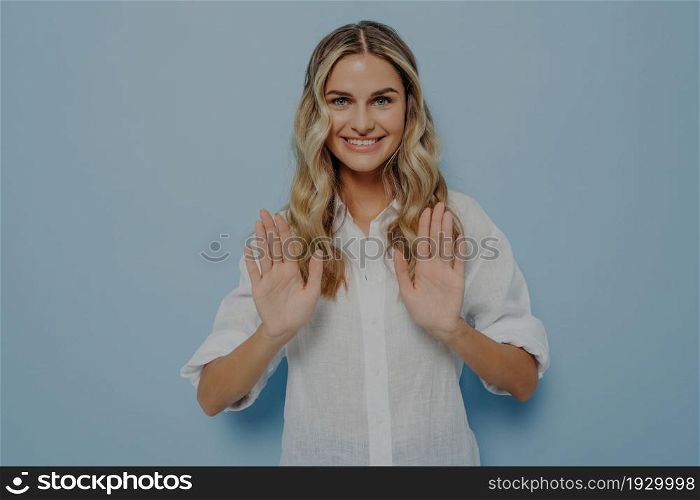 Young woman politely refusing what she is offered, both her hands in front of her, showing her disagreement with gesture while standing alone next to blue wall. Disagreement concept. Blonde teenage girl politely refusing