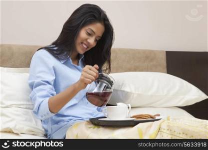 Young woman pouring tea in cup while sitting on bed
