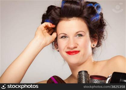 Young woman preparing for date having fun, cute girl with curlers styling hair with many accessories comb brush hairdreyer on gray