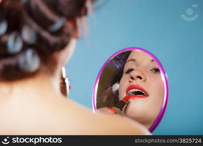 young woman preparing to party, getting ready for going out. Girl styling hair with curlers applying make up red lipstick looking at mirror retro style blue background