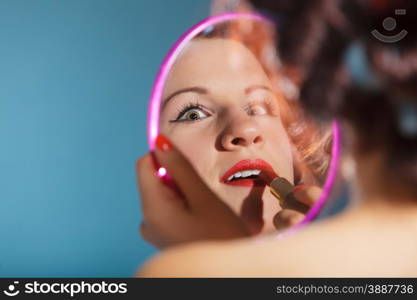 young woman preparing to party, getting ready for going out. Girl styling hair with curlers applying make up red lipstick looking at mirror retro style blue background