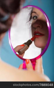 young woman preparing to party, getting ready for going out. Girl styling hair with curlers applying make up red lipstick looking at mirror retro style blue background