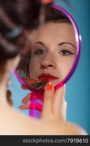 young woman preparing to party, getting ready for going out. Girl styling hair with curlers applying make up red lipstick looking at mirror retro style blue background