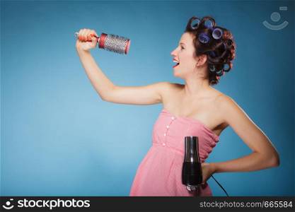 Young woman preparing to party having fun, amusing girl styling hair with curlers hairbrush and hairdreyer retro style blue background