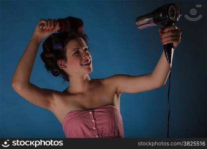 Young woman preparing to party having fun, funny girl styling hair with curlers hairbrush and hairdreyer retro style blue background