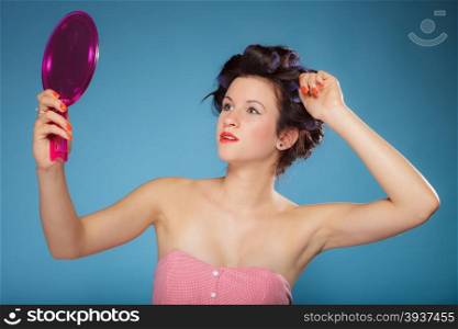 young woman preparing to party having fun, girl styling hair with curlers looking in the mirror retro style blue background