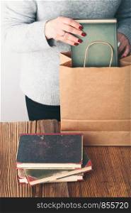 Young woman putting old books to paper bag in antique bookstore. Woman wearing grey sweater and jeans. Vertical photo