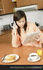 Young woman reading a newspaper and using a mobile phone at a kitchen counter