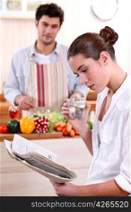Young woman reading a newspaper while her boyfriend prepares lunch