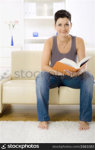 Young woman reading book at home, sitting on sofa.
