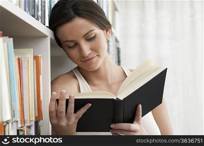Young woman reading book, leaning against bookshelf