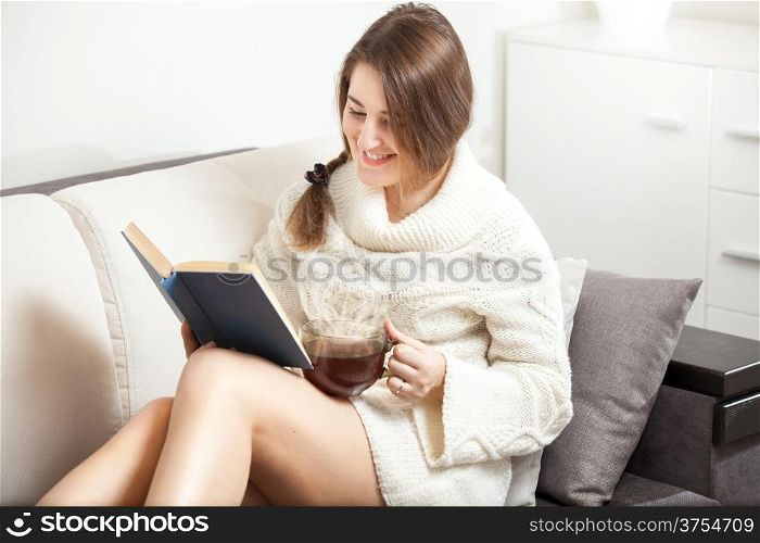 Young woman reading book on sofa and drinking hot tea