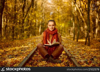 Young woman reading book while sitting railroad track over autumn fallen leaves background. Woman reading book while sitting railroad track