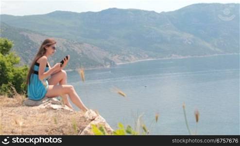 young woman reading e-book while sitting on top of a cliff near the sea. Balaklava, Crimea, Ukraine