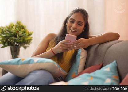 Young woman reading messages on  mobile phone while relaxing on sofa in living room