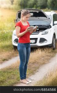 Young woman reading owner manual at broken car in meadow