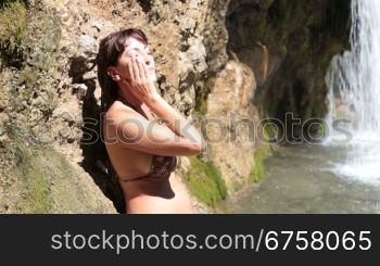Young woman refreshing the face with water from a waterfall on a hot summer day