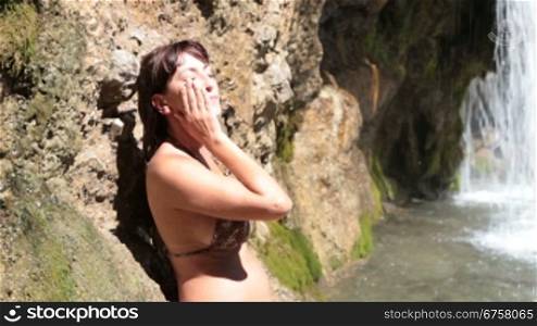 Young woman refreshing the face with water from a waterfall on a hot summer day
