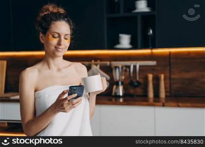 Young woman relaxing at home and reading messages on mobile phone. Happy hispanic girl wrapped in towel applies collagen eye patches after shower. Body care, beauty and wellness. Modern interior.. Happy girl applies collagen eye patches after shower and reading messages on mobile phone.