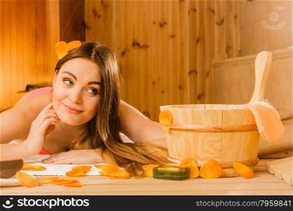 Young woman relaxing in sauna. Spa wellbeing.. Young woman with bucket ladle and petals relaxing laying in wood finnish sauna. Attractive girl in bikini resting. Spa wellbeing pleasure.