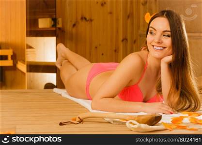 Young woman relaxing in sauna. Spa wellbeing.. Young woman with ladle and petals relaxing laying in wood finnish sauna. Attractive girl in bikini resting. Spa wellbeing pleasure.