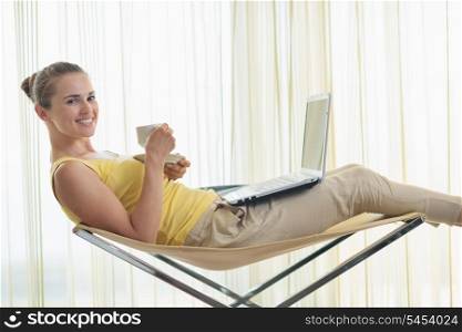 Young woman relaxing on modern chair with laptop and tea