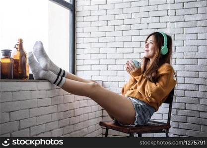 Young Woman Relaxing with Music From Headphone in Cozy House Nearby Window, Enjoying with Morning Sunshine and Hot Coffee