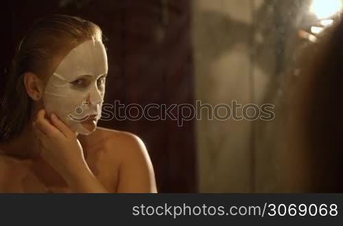 Young woman removing a facial mask and admiring the result looking in the mirror