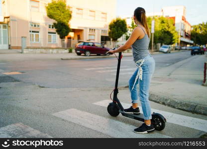 Young woman rides electric kick scooter on the street waiting on the crossroad wearing jeans in summer day evening traffic transport