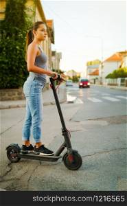 Young woman rides electric kick scooter on the street waiting on the crossroad wearing jeans in summer day evening traffic transport