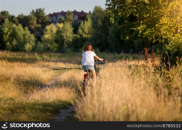 Young woman riding bicycle at meadow on dirt road at sunset