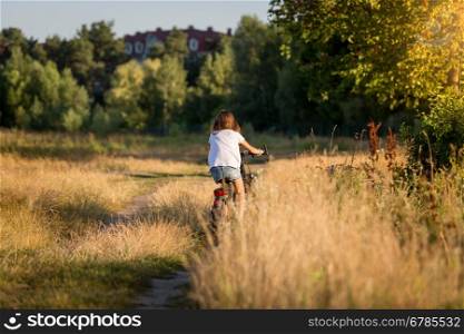 Young woman riding bicycle at meadow on dirt road at sunset