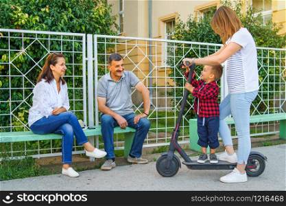 Young woman riding electric kick scooter with her little boy son while talking to grandfather senior man and woman sitting on the bench in summer day