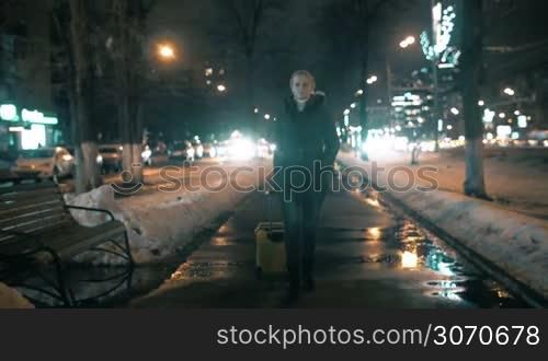 Young woman rolling trolley bag in the city at night. She looks very serious and decisive as if it is way to new life
