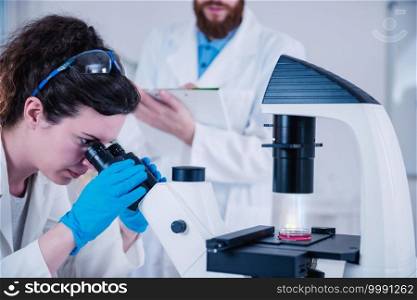 Young woman scientist doing test in laboratory, male scientist taking notes near female colleague working with microscope