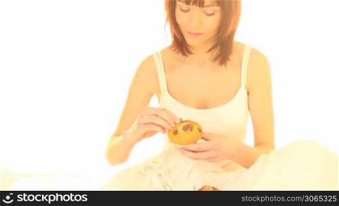 Young woman seated on her bed eating a muffin isolated on white