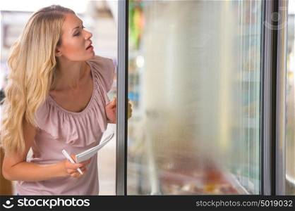 Young woman shopping at supermarket. Holding shopping list and opening fridge to pick up frozen foods