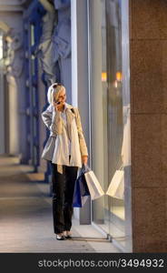 Young woman shopping evening city looking into shop windows phone