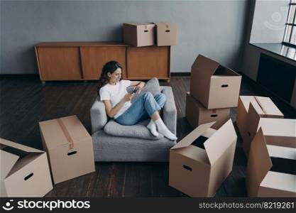 Young woman shopping using online store mobile phone app, purchasing furniture, sitting between carton boxes on moving day. Female choosing relocation service to move packed things to new house.. Woman using phone, purchases furniture, chooses relocation service to move packed things to new home